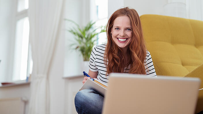 eine rothaarige Frau mit sitzt lächelnd vor einem Laptop mit Block und Stift.