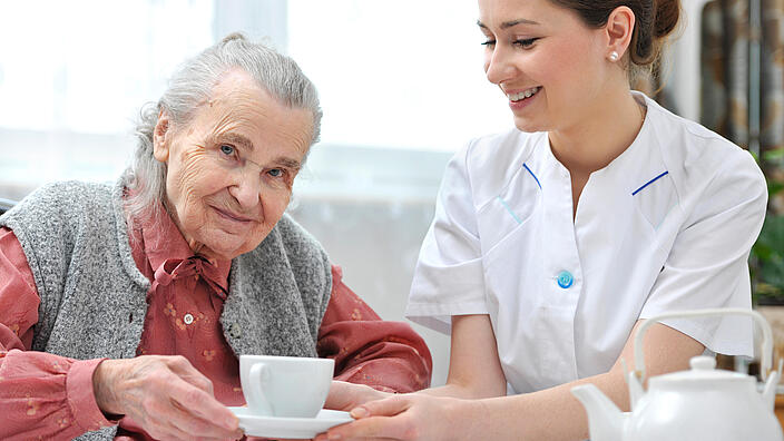 Ältere Frau sitzt neben einer Pflegerin und hält eine Tasse.